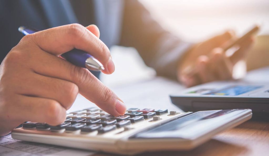 A person using a calculator while holding a pen on one hand and using a phone on another hand.